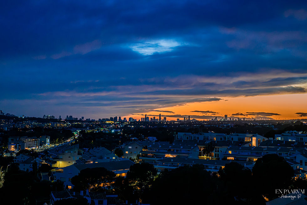 City skyline at dusk with vibrant orange and blue sky. City lights illuminate buildings and streets. Text: En Paarvai - Aishwarya R.