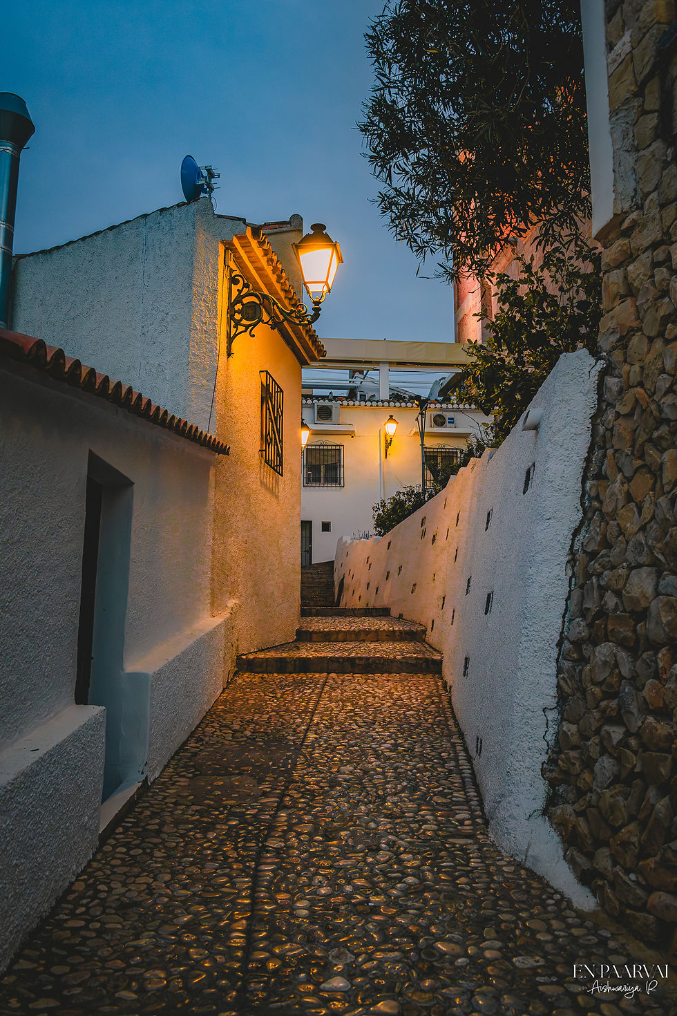 Narrow cobblestone path with white walls, lit by warm street lamps. Evening setting with a calm, inviting atmosphere. Signs of residence visible.