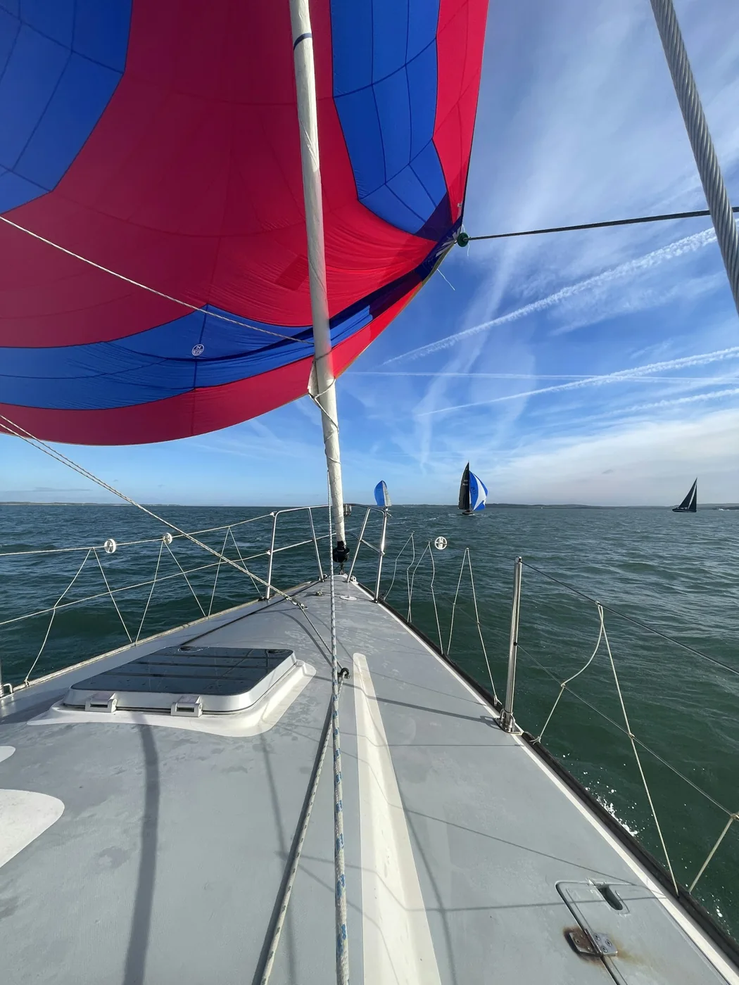 Proa de veleiro sob vela azul e vermelha, com outros veleiros no mar ao fundo.
