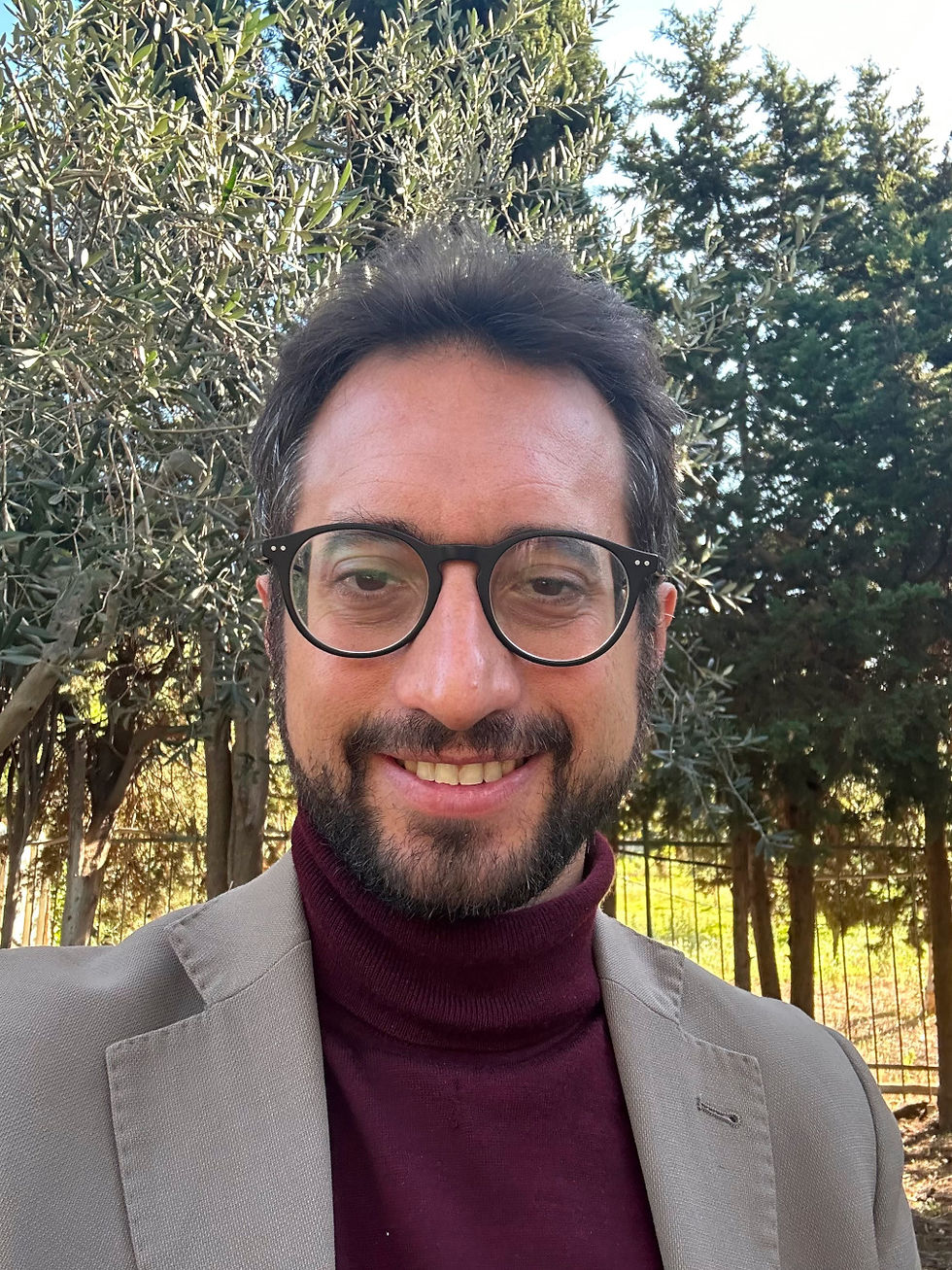 A man stands in an olive grove. He wears glasses and has a black beard and moustache.