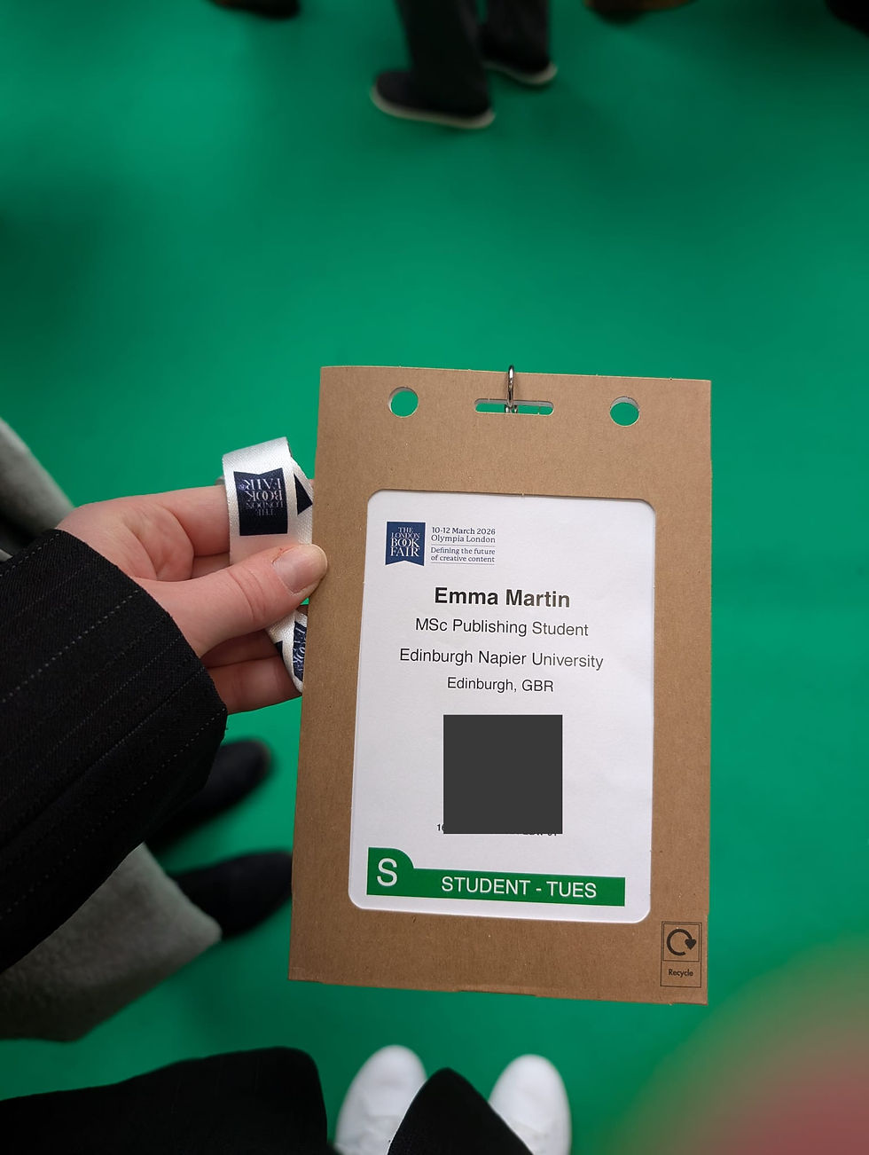 Person holding a name badge for an event at Olympia London, on a green floor background. Badge includes name and university details.