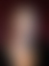 Person wearing glasses and a black top against a dark red background, looking at the camera with a neutral expression.