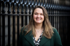 A woman in a green coat smiles in front of a black wrought iron fence. She has long brown hair and a patterned shirt, outdoors.