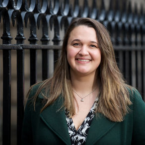 A woman in a green coat smiles in front of a black wrought iron fence. She has long brown hair and a patterned shirt, outdoors.
