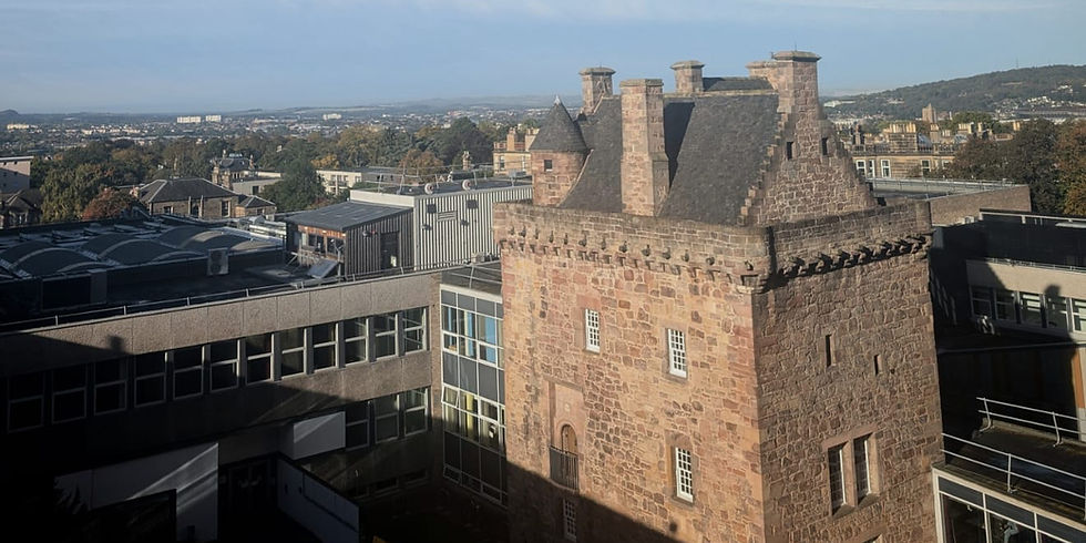 Historic stone tower among modern buildings, with distant hills under a clear blue sky. 