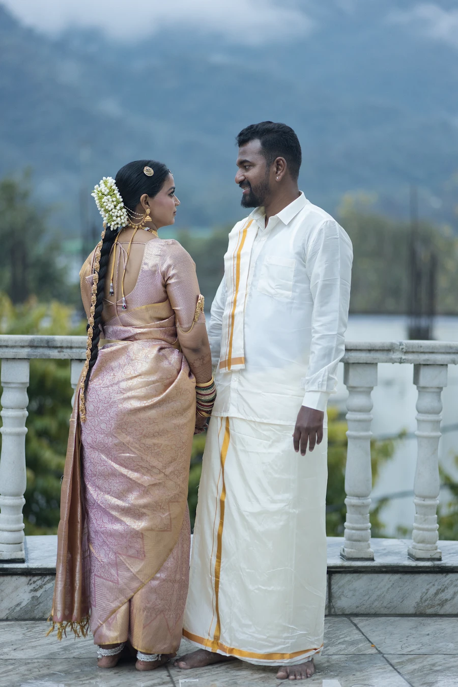 South Indian bride in golden saree with jasmine braid and groom in white mundu smiling at each other during destination wedding shoot in the hills