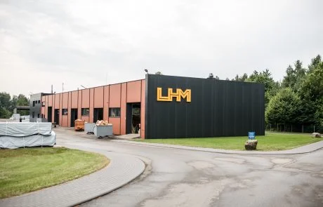 Wide landscape photo of the LHM industrial building exterior with logo on the dark facade, overcast sky, driveway and green lawn.