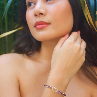 Woman wearing a heart-shaped bracelet on wrist, looking right, tropical background.