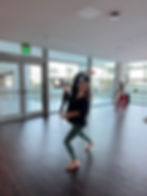 Women dancing in a bright studio with large windows. The leader in green leggings smiles while leading others. Exit sign visible. Calm mood.