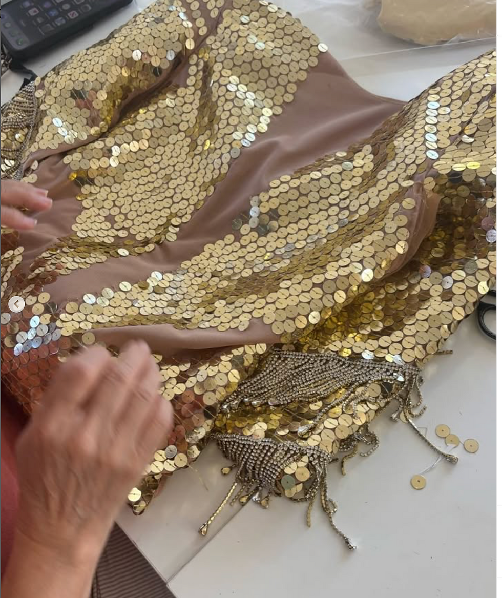 Hands sewing gold sequins onto beige fabric with fringe details. Phone nearby on table. Creative and focused setting.