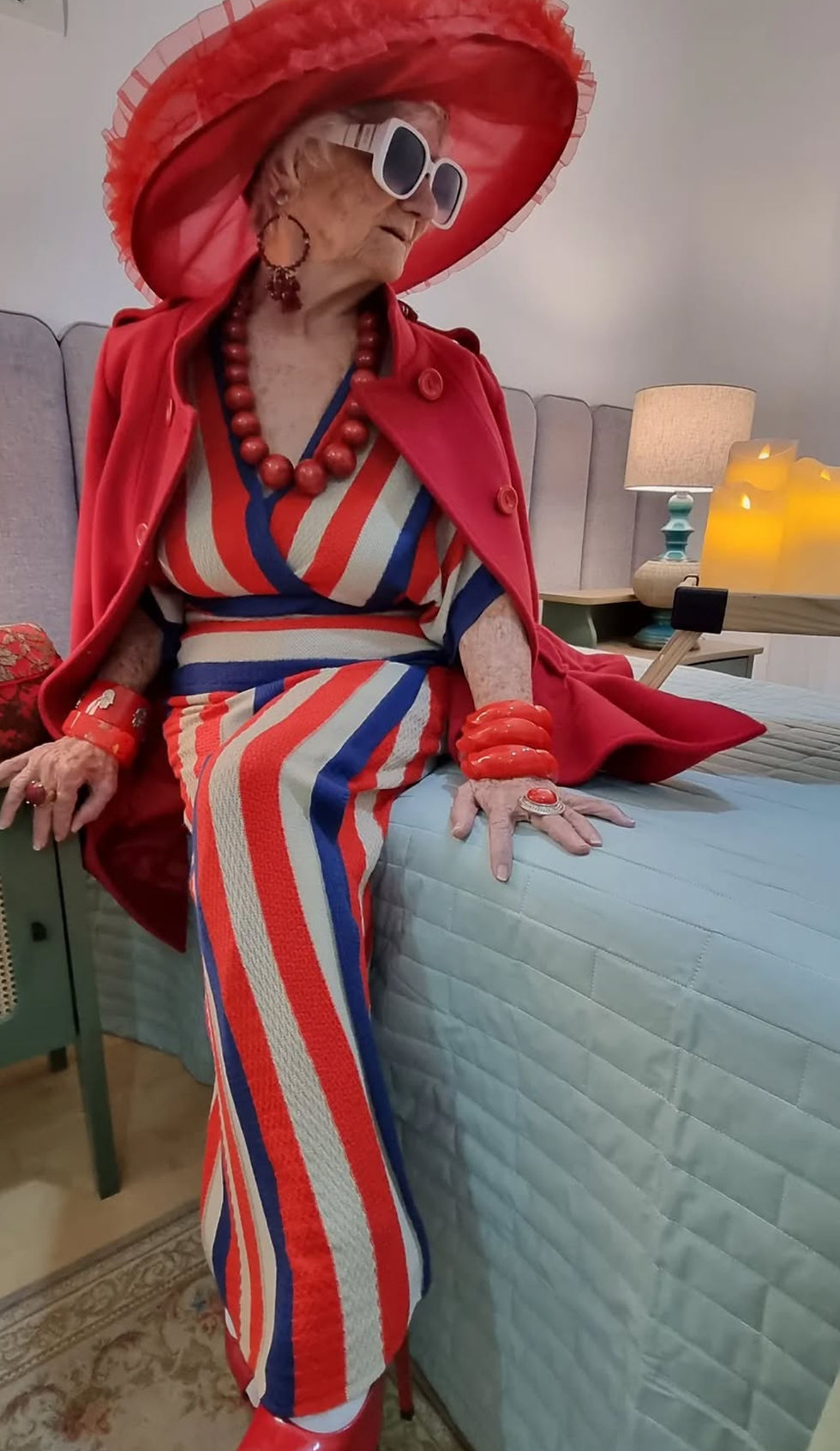 Vovó Izaura Demari.n bold red and blue outfit, large hat, and sunglasses. Sits confidently indoors on a bed next to a lit lamp.