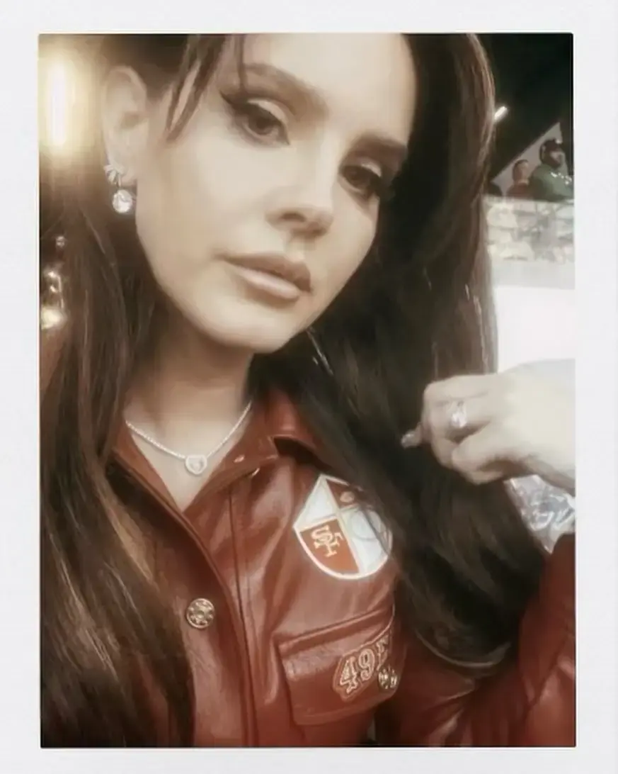 Lana Doe Rey in in a custom sugar ferrini red jacket with patches, adjusting hair. Background is blurred. She has a neutral expression, wearing earrings and a necklace.