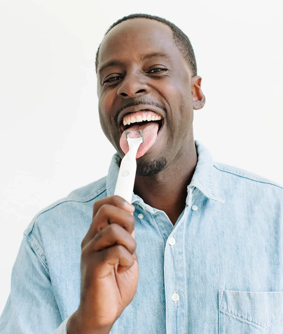 Man in a denim shirt using a white tongue cleaner, smiling in a bright setting, conveying a fresh and clean feeling.