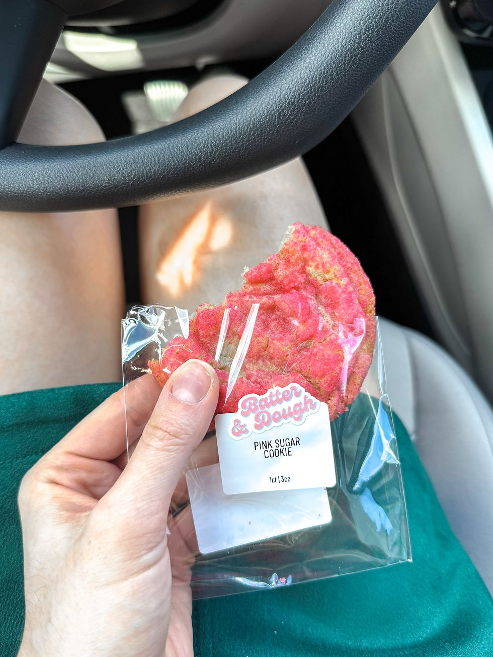 Hand holds a bitten pink sugar cookie in a package labeled "Batter & Dough" inside a car, with steering wheel and green skirt visible.