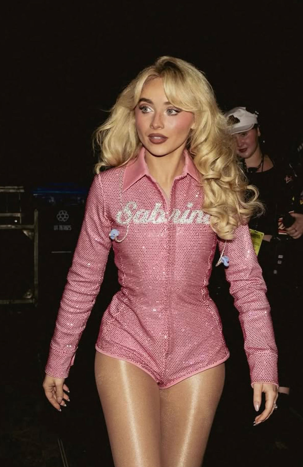 Sabrina Carpenter in pink sequined Sugar Ferreni outfit with "Sabrina" text, walking confidently. Dark background with hints of people and equipment. Mood: Glamorous.