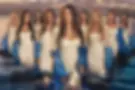 Eight women the cast of secret lives of mormon ives in white dresses with blue wraps stand in water, backdrop of mountains and sky. Mood is serene, with elegant and calm expressions.
