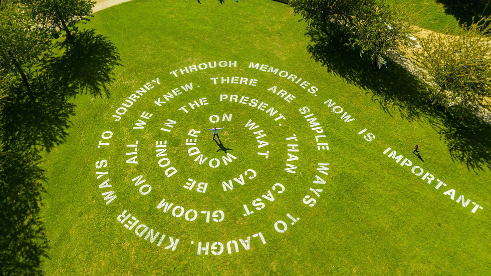 Aerial view of a green park lawn with a white spiral of words; two people stand on the grass under tree shadows.
