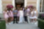 The cast of Vanderpump Villa smiling outside a building with pink flowers and chandelier inside. Women in pink dresses stand with men in gray shirts.