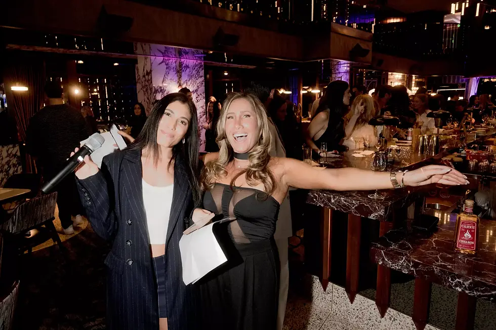 Two women smiling at a lively bar, one holds a microphone. The dim setting has purple lighting and marble counters. Guests mingle nearby.