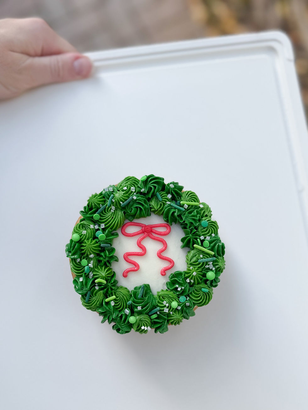 Hand holding a white board with a cupcake decorated as a green wreath with a red frosting bow. Background is blurred.
