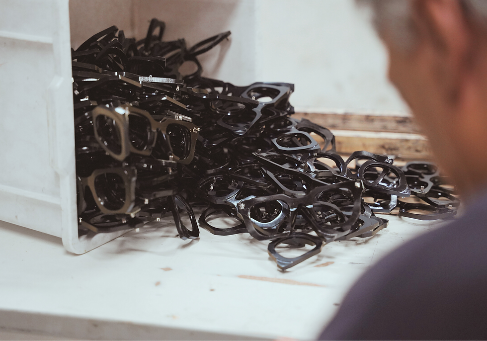 A pile of black eyeglass frames next to a white bin on a workbench. A person is partially visible, focused on the frames.