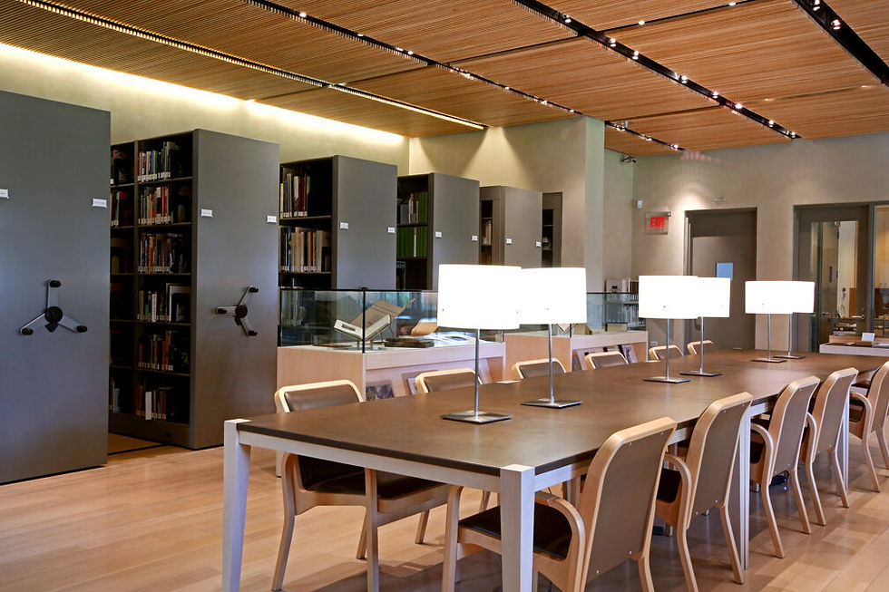 Modern library reading room with long wooden table, chairs, glowing lamps, bookshelves, and a display case under a slatted ceiling.