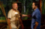 Two men in a tribal setting; one extinguishes a torch held by the other, evoking a somber mood. Dense foliage in the background.