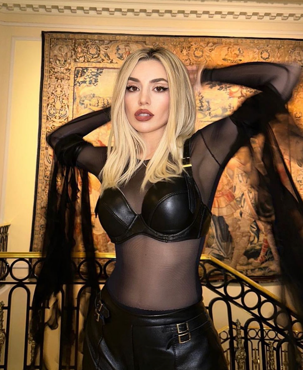 Ava Max in custom Sugar Ferini black leather and sheer top poses with arms raised. Ornate tapestry and black railing in background, creating a stylish vibe.