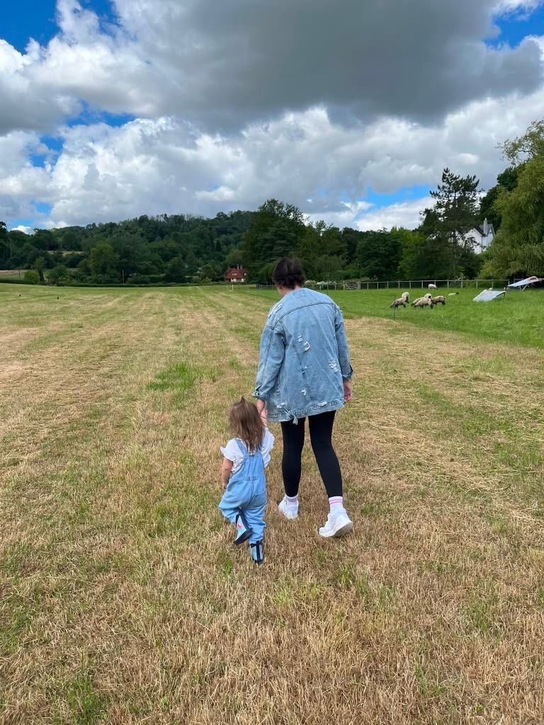 Woman and toddler walk through a grassy field toward grazing sheep under a cloudy sky.