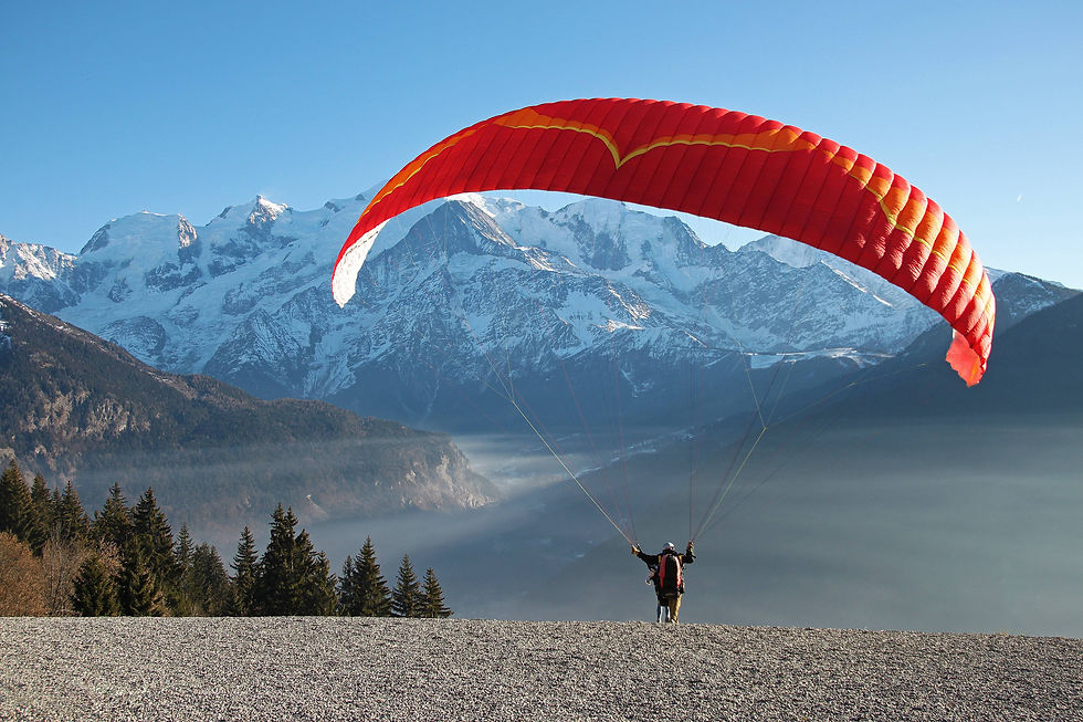 Person paragliding with a red wing over foggy mountain landscape. Snow-capped peaks and evergreen trees in the background, clear sky above.