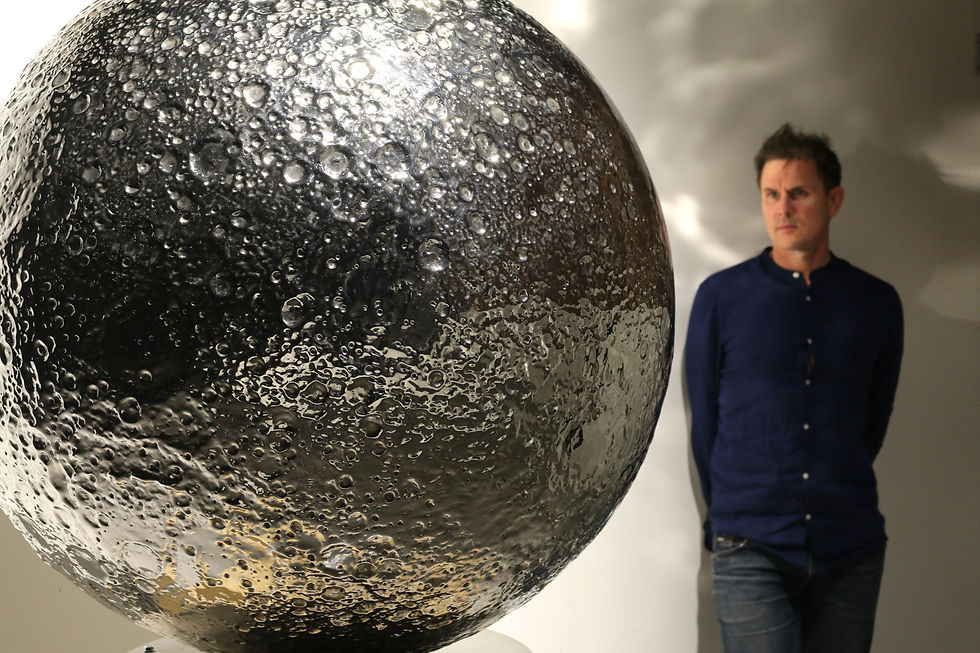Large cratered metallic sphere sculpture beside a man in a gallery, with soft light and a serious mood.