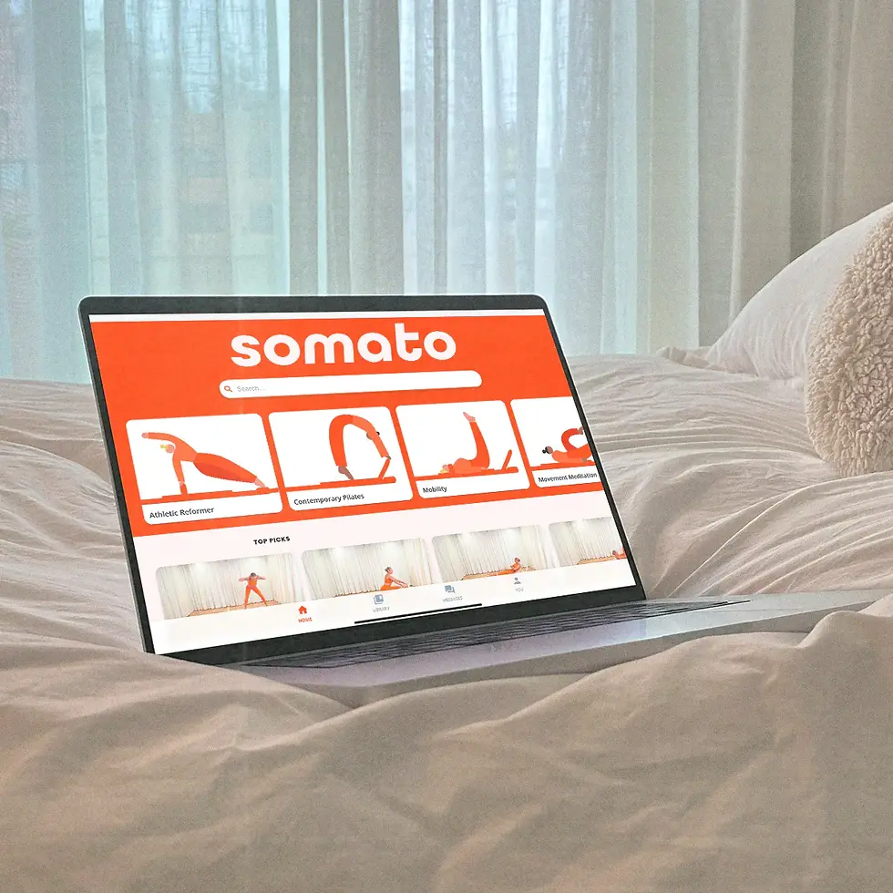 Laptop on a bed, displaying a fitness website with an orange header reading "somato." The room has light curtains and a cozy ambiance.