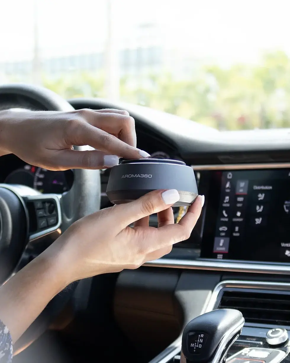Hands adjusting an Aroma360 diffuser inside a car, with a touchscreen display and gear shift visible. Bright, sunny background.