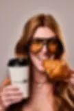 Woman with long hair, wearing large brown sunglasses, smiles while holding a croissant and coffee cup labeled "Make Me Drip." Neutral background.