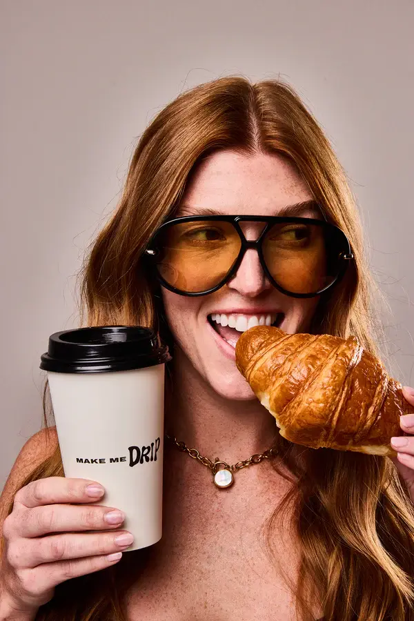 Woman with long hair, wearing large brown sunglasses, smiles while holding a croissant and coffee cup labeled "Make Me Drip." Neutral background.