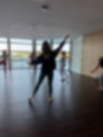 People in a dance class stretch in a bright studio with large windows. The floor is wooden, and participants wear colorful activewear. Mood: focused.