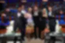 Six people in formal attire pose smiling with arms open on a TV show set. A cheering audience sits in the colorful background.