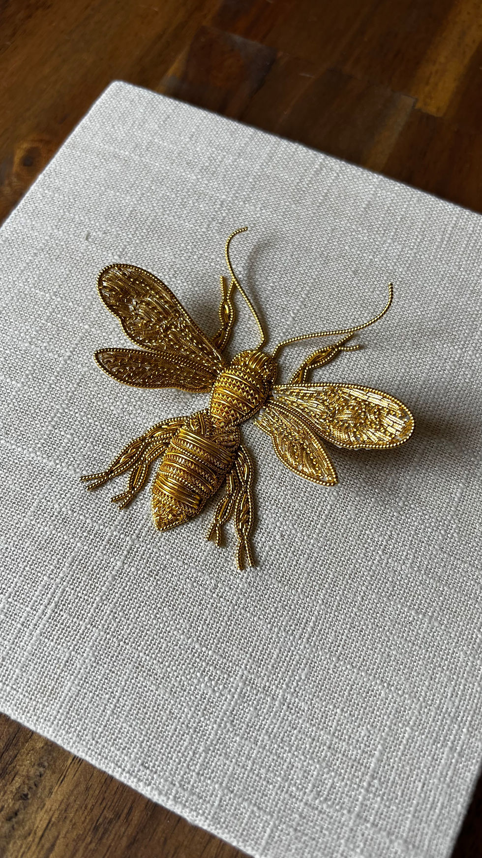 Gold bee-shaped brooch with filigree wings resting on a white textured cloth on a wooden table.