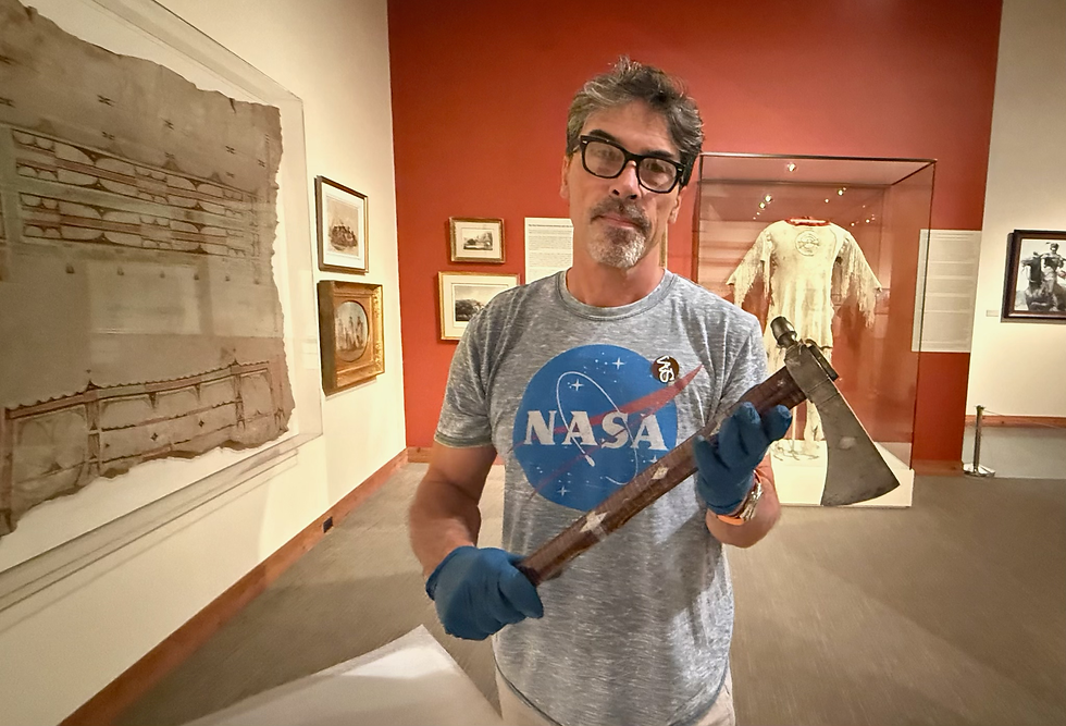 Denis Belliveau in a NASA shirt holds a tomahawk in a museum with framed artifacts and a fringed garment in the background. He wears blue gloves.