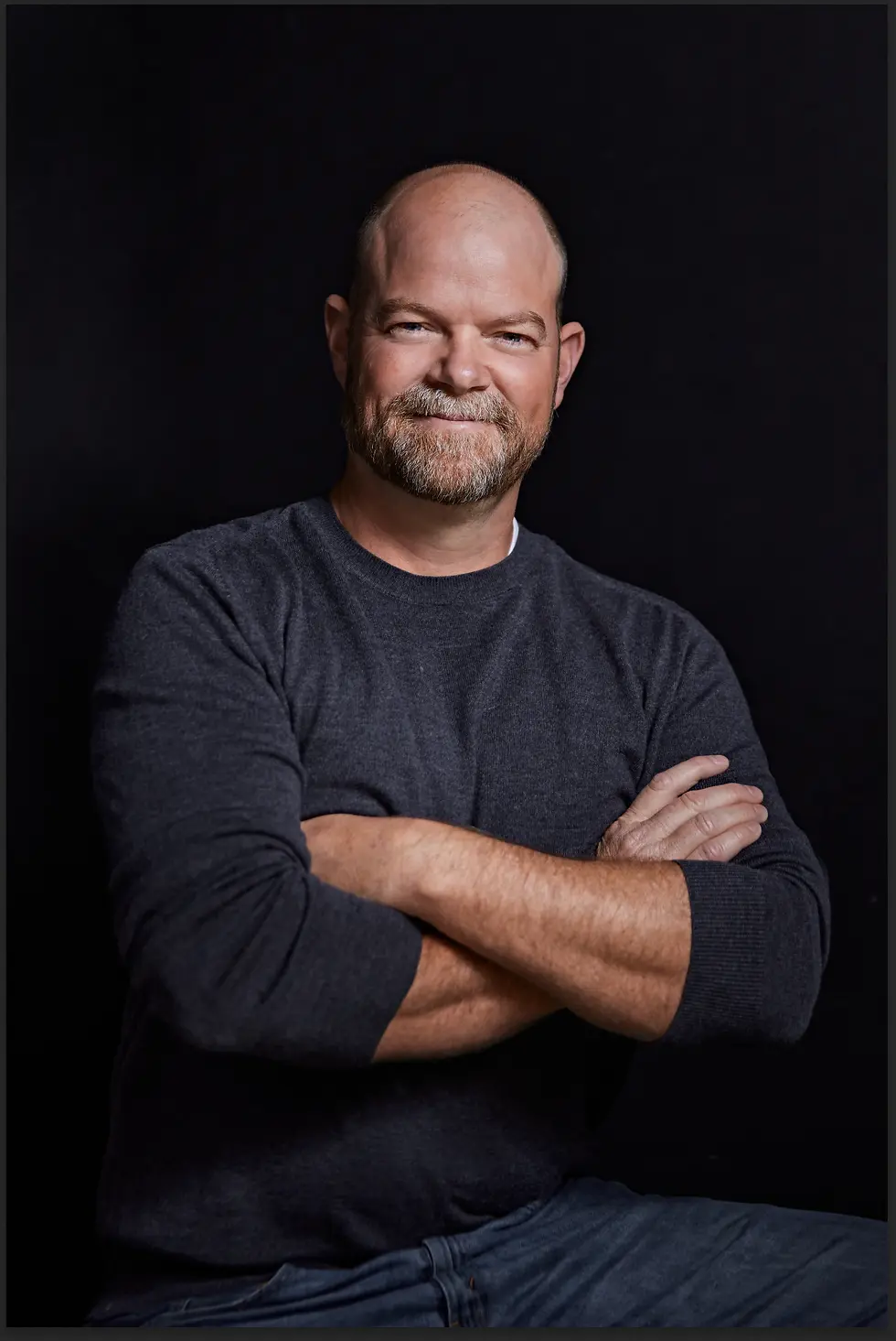 Bald man with a beard, smiling, arms crossed, wearing a dark sweater against a black background. Mood is confident and relaxed.