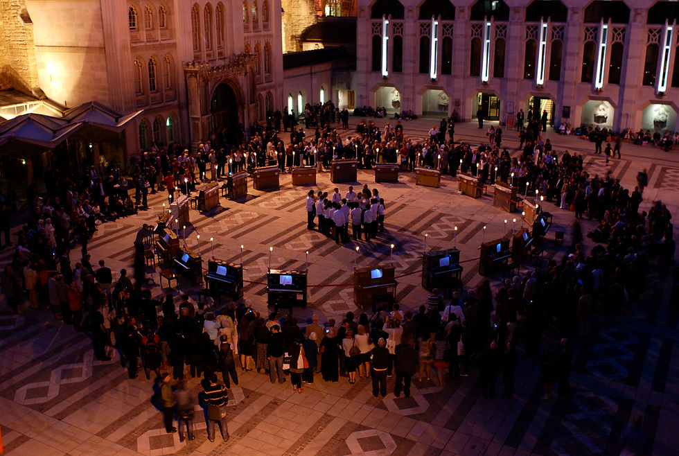 Crowd gathers in a lit stone courtyard at night around a ring of pianos and candlelights, watching a live musical performance.