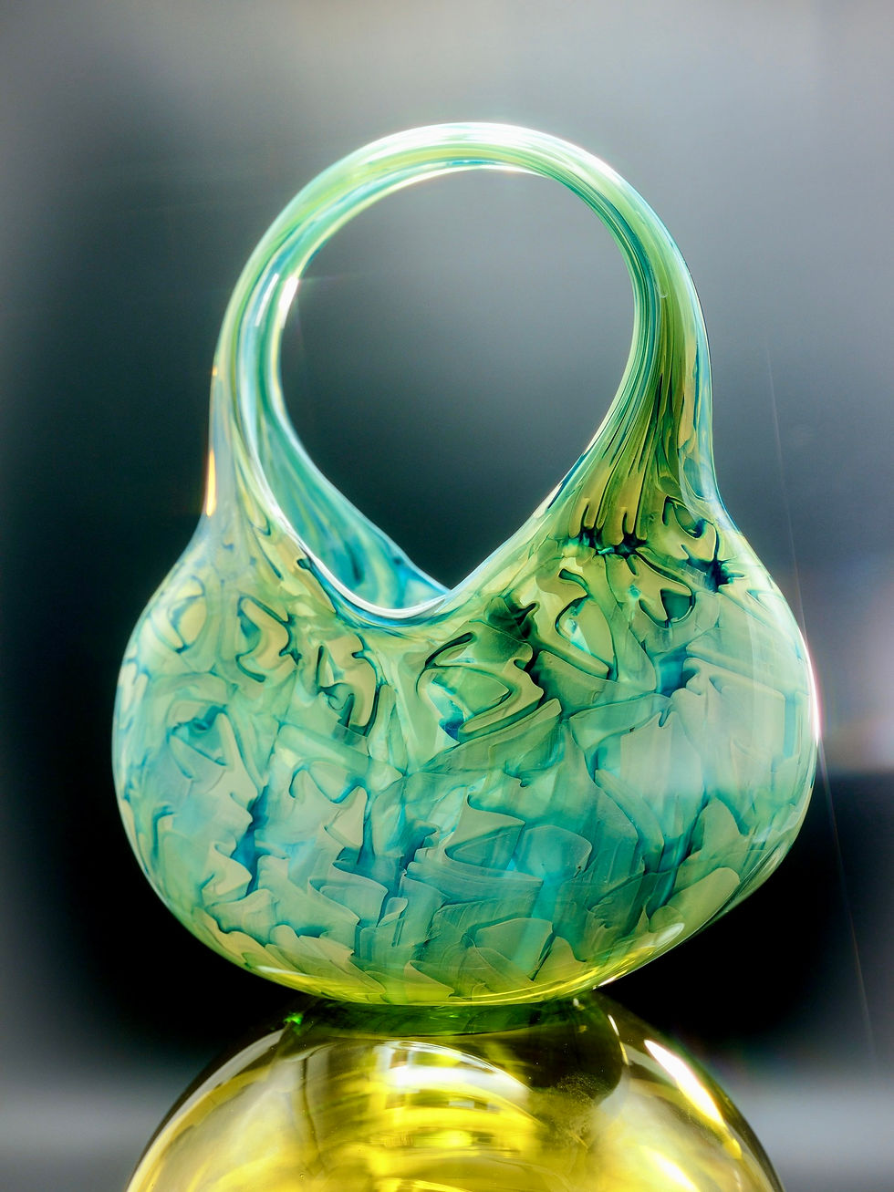 Colorful glass sculpture in teal and green hues, resembling a basket, with swirling patterns against a soft gray background.