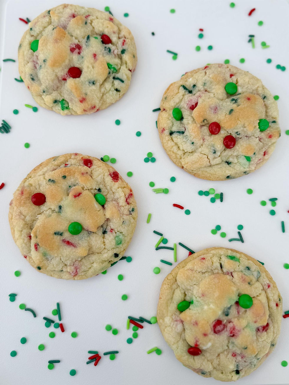 Four colorful cookies with red and green sprinkles sit on a white surface, surrounded by scattered matching sprinkles. Festive and bright.