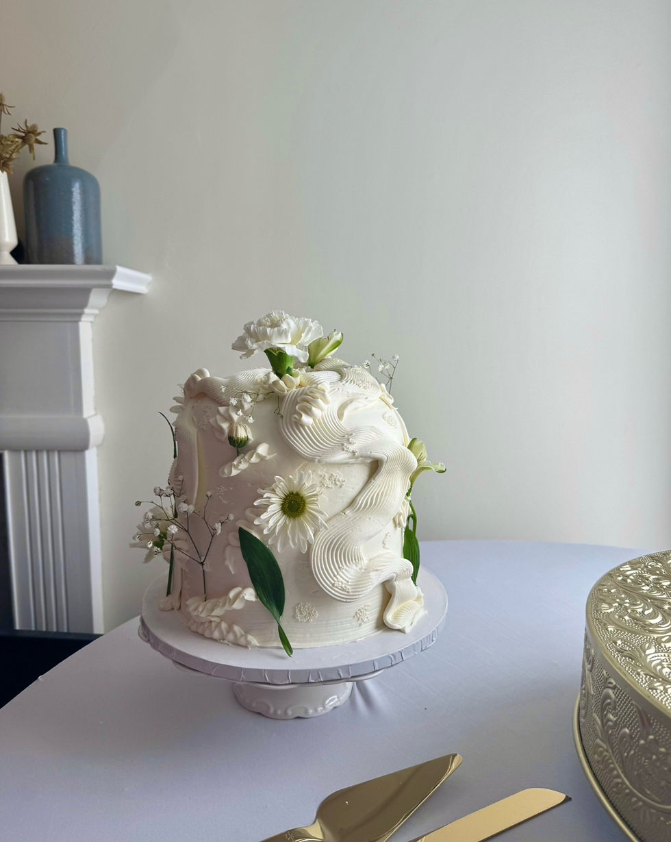Elaborately decorated white cake with flowers on a stand, set against a white wall. Nearby are gold utensils and a patterned container.