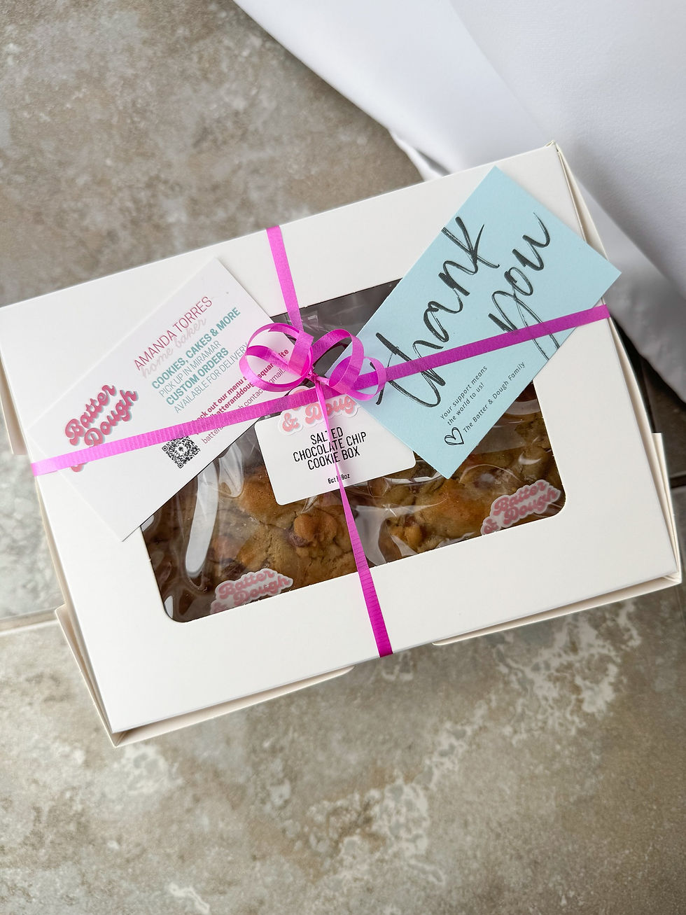 White pastry box with cookies, tied with a pink ribbon. Blue "thank you" card and business card on top. Gray textured floor background.