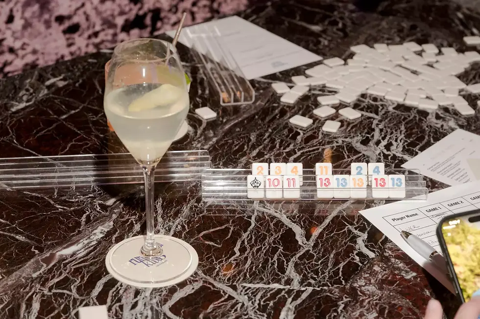 Marble table with Rummikub tiles, glass of cocktail, shuffled tiles, and score sheet. Hand writing on paper, smartphone partially visible.
