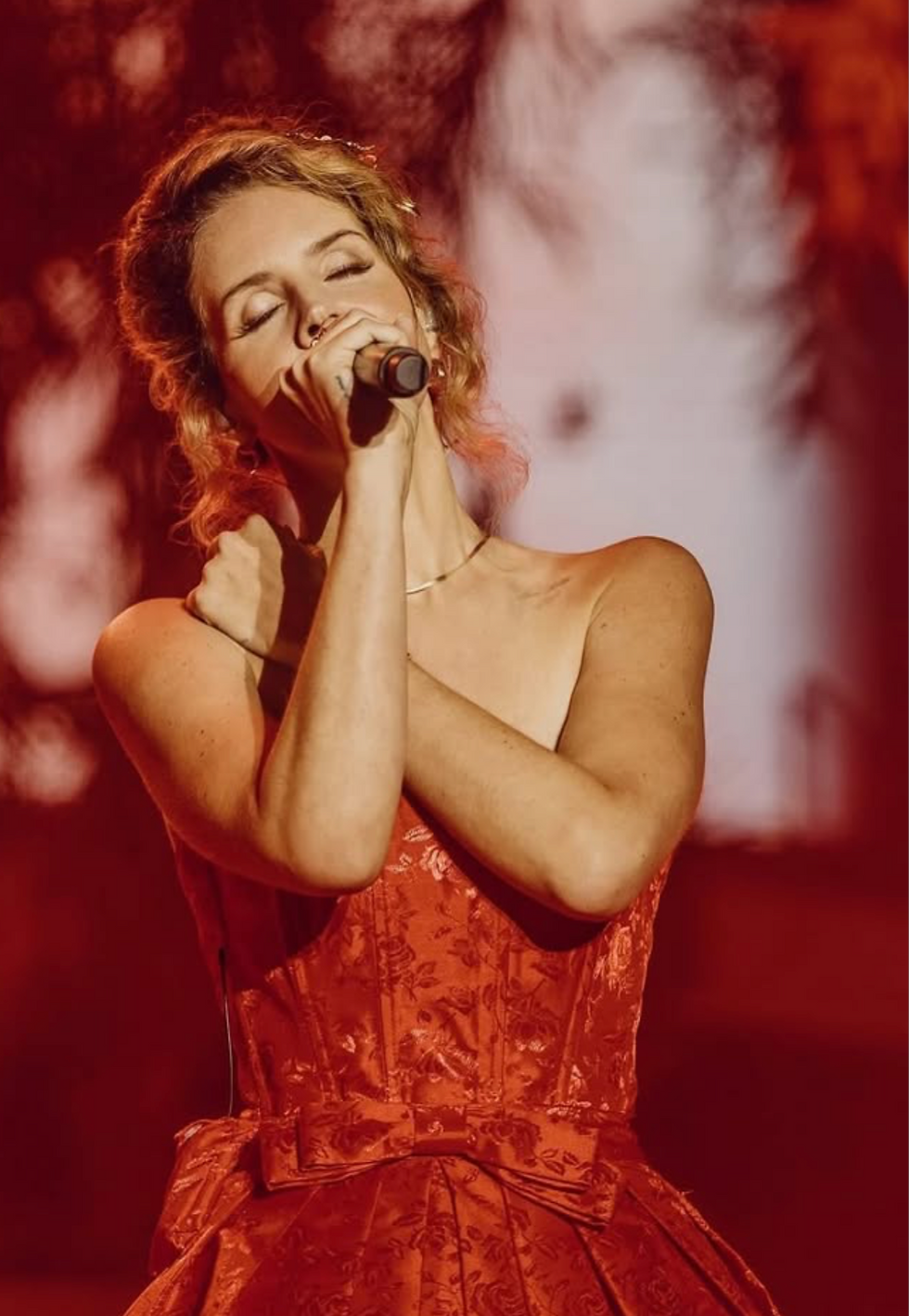 Lana Del Rey in a Sugar Ferrini red dress with eyes closed, holding a microphone on a red-lit stage, evoking an emotional, intimate performance.