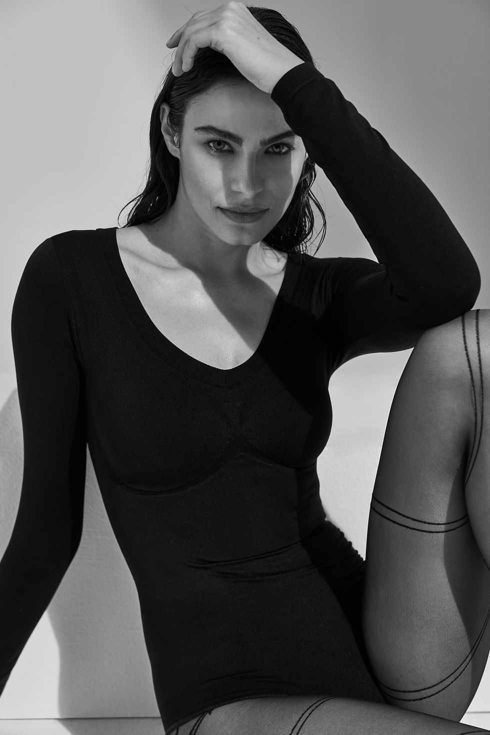 Black-and-white portrait of a woman in a black bodysuit, hand on head, seated against a plain backdrop, intense gaze.