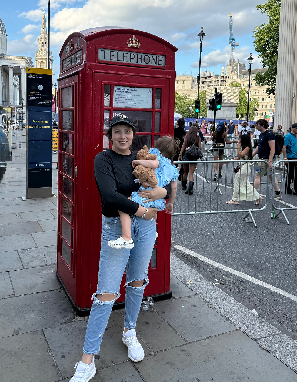 Smiling woman holds a toddler with a teddy bear beside a red London telephone booth on a busy street, with Trafalgar Square behind.