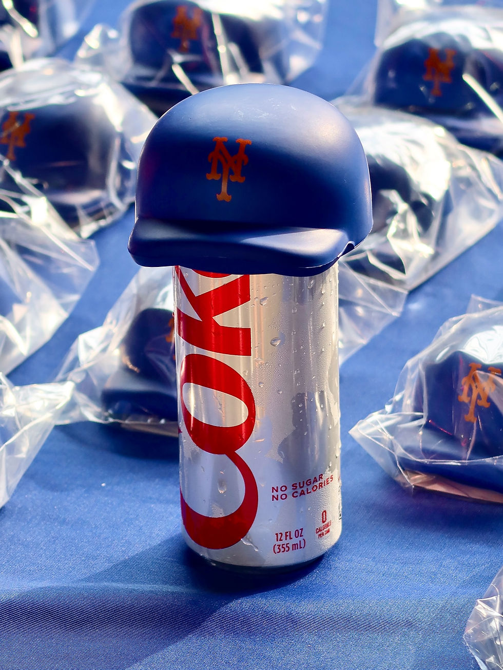 Diet Coke can with a small blue sports helmet on top featuring an orange logo. Background has more helmets wrapped in plastic, set on a blue surface.